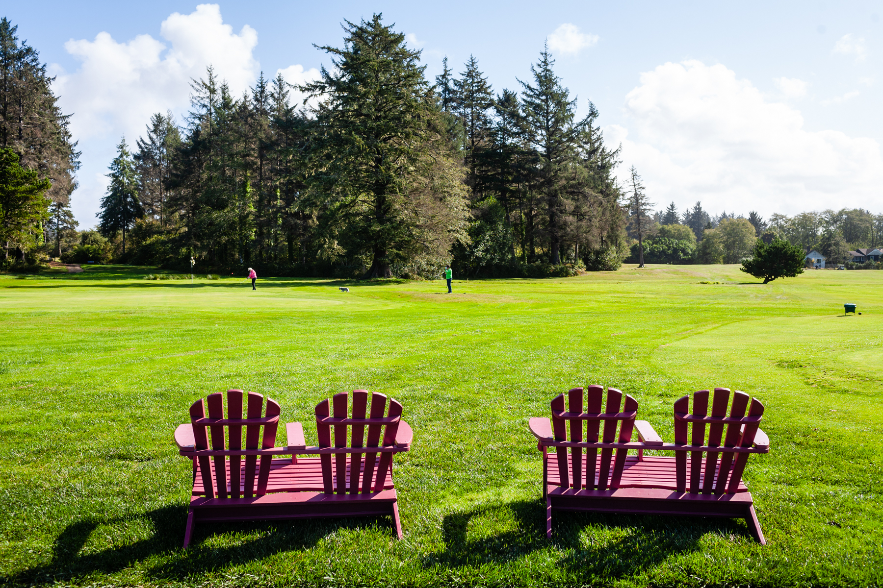 Two Adirondack chairs overlook a lush green at Peninsula Golf Course with golfers and a dog enjoying the course in the distance